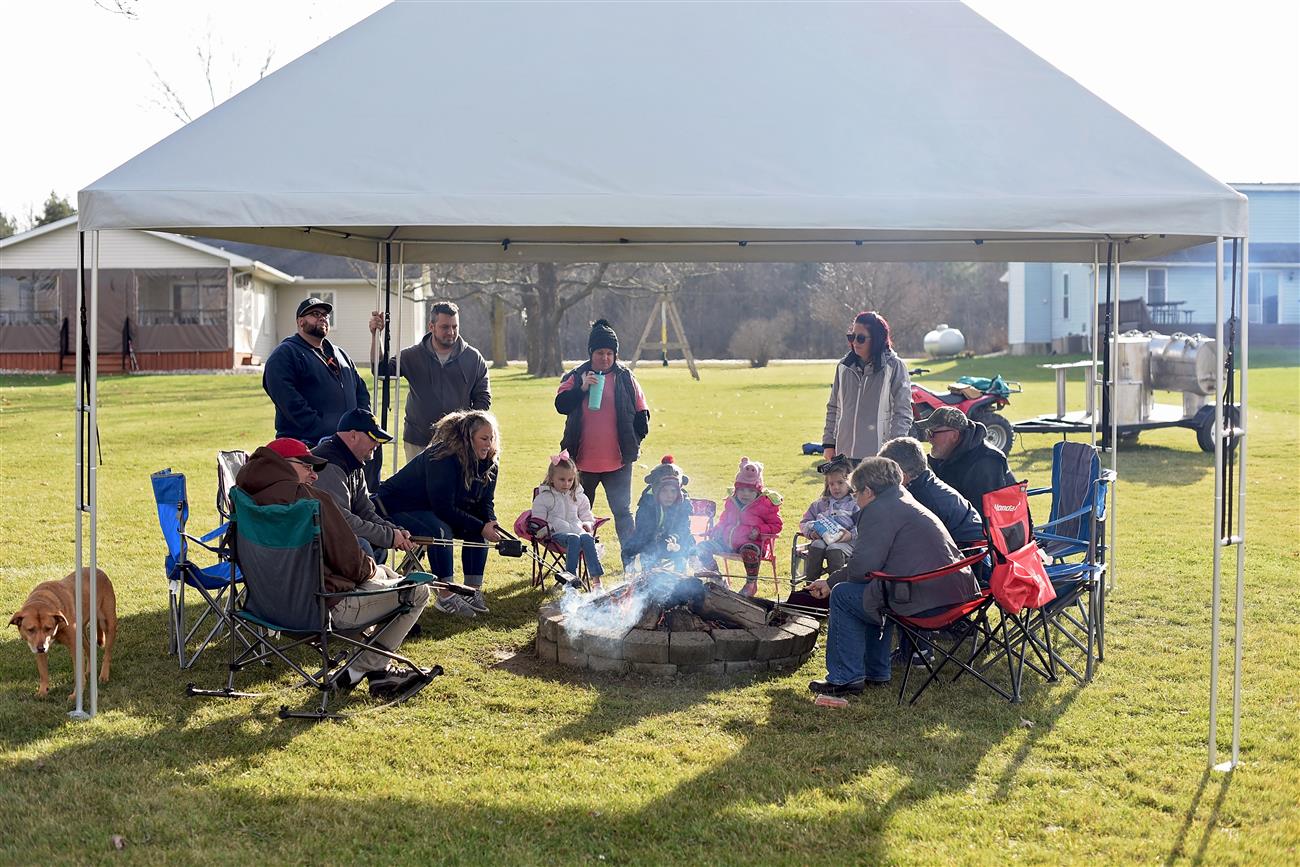 A group of people gathered around a campfire on a lawn, under a large canopy, with a dog, chairs, and some homes in the background.